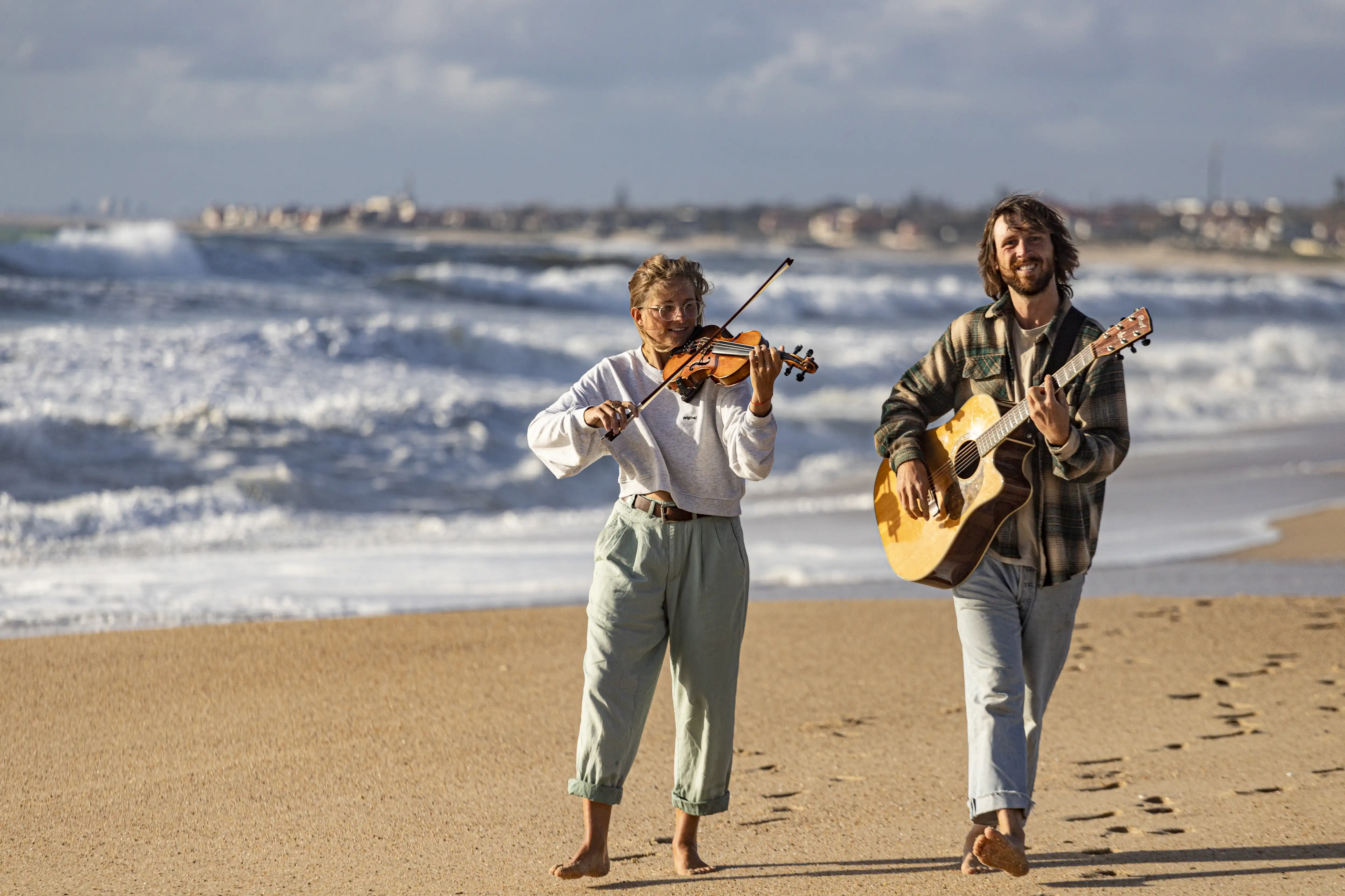 Anka und René laufen am Strand und spielen auf der Geige und auf der Gitarre.
        Die Sonne scheint und im Hintergrund brechen weiße Wellen.